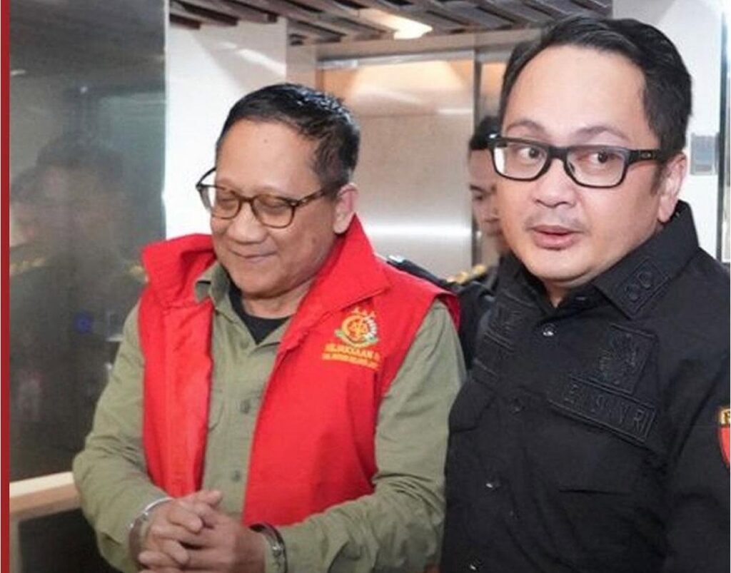 Two men stand indoors a man in a red vest with an embroidered crest and green shirt has his hands clasped while a man in a black security uniform with glasses stands beside him